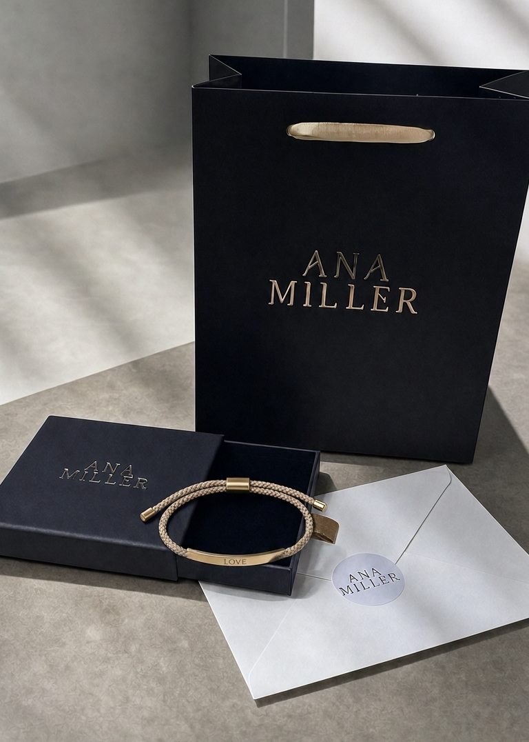 A gold-tone metal bracelet with a textured woven band, displayed next to its branded black presentation box and a white envelope on a gray surface. A black shopping bag with gold 'ANA MILLER' lettering stands in the background.